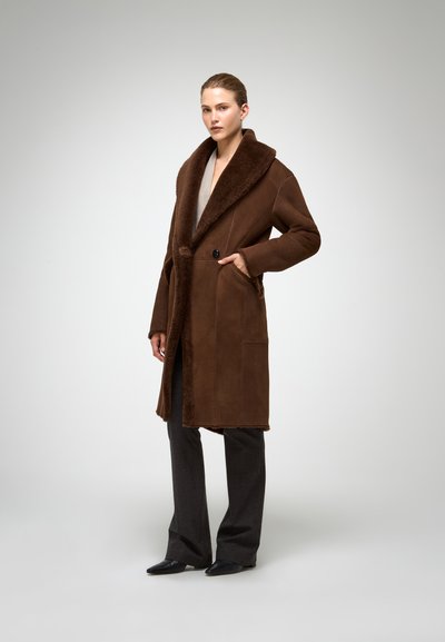 Brūna šūtu jaka ar plašu, struktūrētu apkakli. Aprīkota ar vienu pogu un sānu kabatām, papildināta ar taisna griezuma tumšiem biksēm.