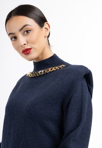 Pulover tricotat bleumarin cu guler înalt, având un accent de lanț aurit la guler și un material texturat.