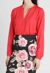 Donna che indossa una blusa rossa a maniche lunghe infilata in una gonna nera con grandi motivi floreali di rose rosa e farfalle.