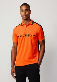 Oranžové polo tričko s černým a olivově zeleným designem, na přední straně s nápisem "HACKETT" v černé barvě. Krátký rukáv.