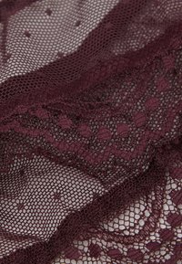 Tissu en dentelle bordeaux présentant des motifs intriqués avec des pois et des bords festonnés, mettant en valeur une texture délicate et semi-transparente.