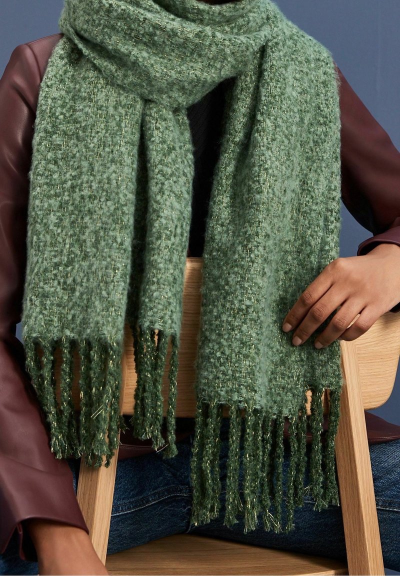 Personne portant une écharpe verte texturée avec des franges, un haut sombre, une veste en cuir bordeaux et un jean bleu, assise sur une chaise en bois.