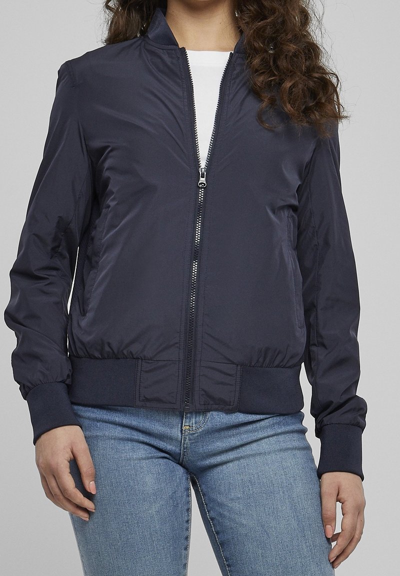 Personne portant une veste bomber bleu marine avec une fermeture éclair devant, un haut blanc et un jean bleu clair, debout devant un fond uni.