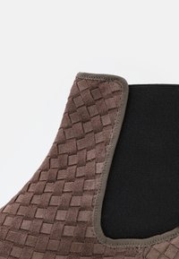 Texture en daim marron avec un motif tissé, section élastique noire et une finition foncée. Accent sur le mélange de matériaux et les éléments de design cohérents.