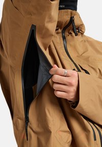 Brązowa kurtka z odsłoniętym czarnym zamkiem błyskawicznym rozpinanym przy bocznym rozcięciu przez rękę noszącą srebrny pierścionek.