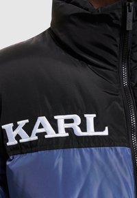 Vadderad jacka med hög krage, svart övre del och blå nedre del. Broderad "KARL"-logotyp i vitt och framdragkedja.