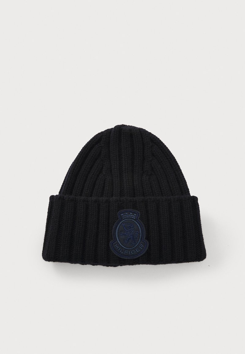 Beanie a coste nera con orlo ripiegato. Presenta un'etichetta ricamata blu scuro con un design di leone. Texture morbida.