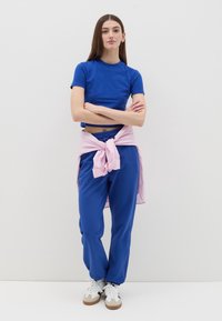 Blauwe katoenen cropped t-shirt en blauwe jogger broek, met een roze gestreepte shirt geknoopt om de taille. Witte sneakers met lichtbruine zolen.