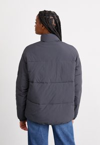 Veste matelassée grise avec col montant, vue de dos. Présente des sections matelassées et des poignets élastiques, fabriquée en tissu léger et lisse.