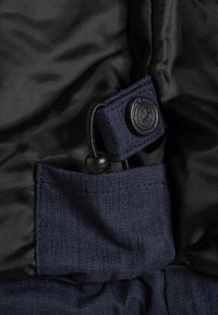 Dunkelblaues Stofftaschen mit strukturierter Oberfläche, ausgestattet mit einem schwarzen Knopfverschluss und verstellbarem Kordelzug, auf schwarzem Innenfutter.