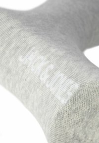 Šedá bavlněná ponožka s jemnou texturou a bílým logem. Materiál je hladký a elastický, navržený pro pohodlí a dokonalé padnutí.