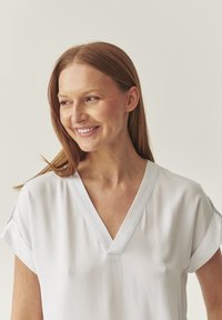 Blusa blanca de manga corta con escote en V. Tejido suave, corte relajado, brillo sutil y mangas dobladas. Diseño minimalista, sin estampados.