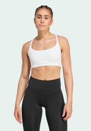 Femme en forme avec des cheveux tressés portant un soutien-gorge de sport blanc et des leggings noirs taille haute sur un fond clair.