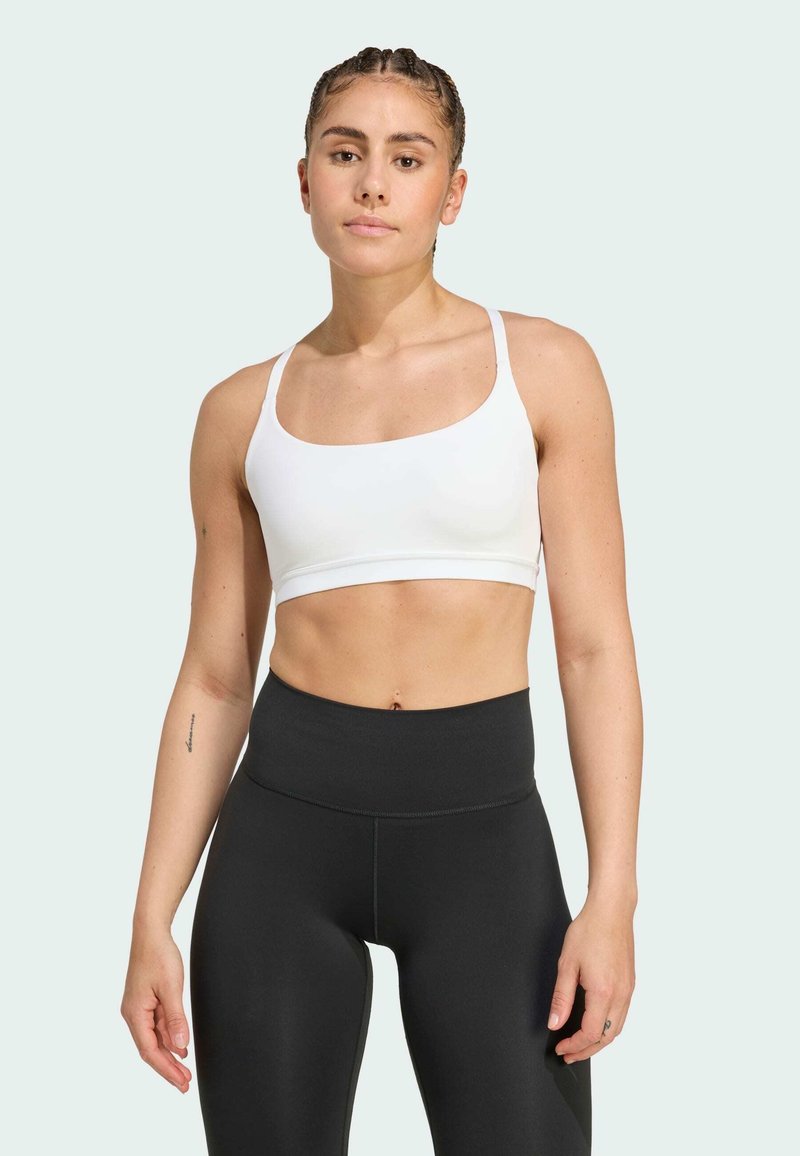 Femme en forme avec des cheveux tressés portant un soutien-gorge de sport blanc et des leggings noirs taille haute sur un fond clair.