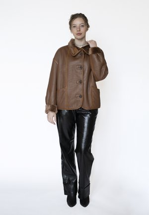 Vrouw die een bruine shearling jas met grote knopen en zwarte glanzende leren broek draagt, staand tegen een witte achtergrond.