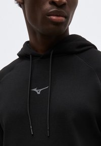 Detailní záběr osoby v černé mikině s kapucí se šňůrkami a malým bílým logem Mizuno na hrudi.