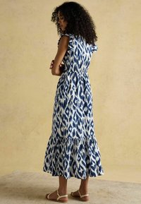 Zila un balta rakstaina maksi kleita ar viļņotām piedurknēm, elastīgu vidukli un pakāpenisku svārku, apvienota ar neitrālas krāsas sandāliem.