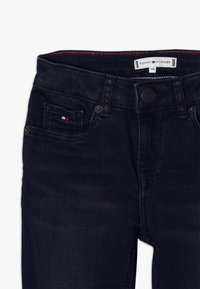 Vaqueros de mezclilla azul oscura con un ajuste slim, diseño de cinco bolsillos, cierre de botón metálico y etiqueta de Tommy Hilfiger en la cinturilla.