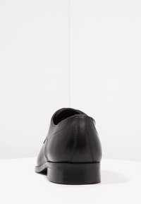 Chaussure habillée en cuir noir vue de dos, montrant les détails des coutures et un petit talon empilé sur une surface blanche.