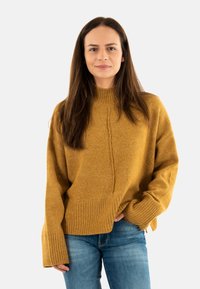 Pull à col montant de couleur moutarde, avec des poignets et un ourlet côtelés. Présente une couture frontale texturée et un design surdimensionné. Associé à un jean bleu.