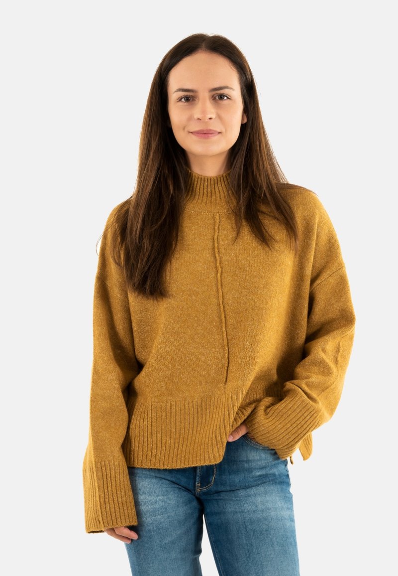 Pull à col montant de couleur moutarde, avec des poignets et un ourlet côtelés. Présente une couture frontale texturée et un design surdimensionné. Associé à un jean bleu.