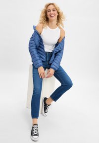 Hellblaues Pufferjacke, enges weißes Tanktop, dunkelblaue Jeans, schwarze Leinensneaker mit weißen Sohlen, sitzende Pose auf einem Podest.
