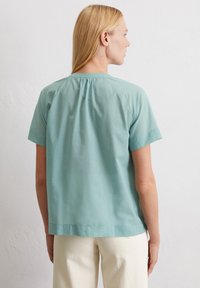 Blusa a maniche corte di un leggero blu verdastro con scollo rotondo, retro arricciato e texture liscia, abbinata a pantaloni color crema.