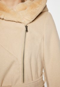 Béžová fleecová bunda s kapucí podšitou umělou kožešinou, stříbrným zipem a páskem v pase. Měkká textura a viditelné ostré designové detaily.