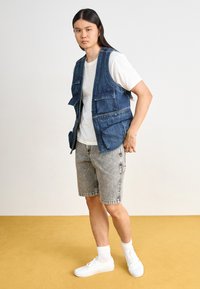 Denim vest with multiple pockets, worn over a white t-shirt. Paired with gray shorts and white sneakers. Standing on a yellow carpet.