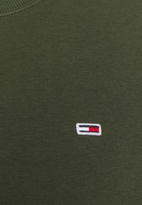 Olivgrön tyg med en liten rektangulär broderad logotyp i rött, vitt och marinblått nära halsringningen.