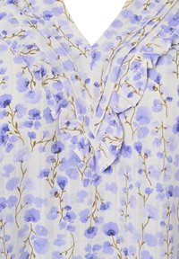 Blouse fleurie en tissu léger avec un col en V orné de volants. Fleurs violettes et tiges vertes sur fond blanc.