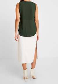 Blusa sin mangas de color verde oscuro con dobladillo redondeado, combinada con una falda midi blanca con una abertura lateral. La modelo lleva botas beige de tacón alto.