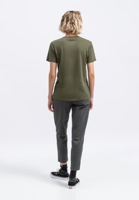 Olijfgroen t-shirt met een ronde hals en korte mouwen, gecombineerd met grijze tapered pants. Model staat met de rug naar de camera, wat een casual outfit laat zien.