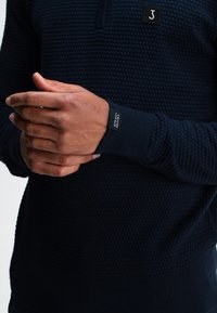 Navyblauwe gebreide trui met een hoge kraag, voorzien van een merketiket op de borst en geribbelde manchetten. Het materiaal lijkt zacht en warm te zijn.