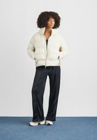 Kobieta o kręconych włosach, ubrana w jasnobeżową kurtkę puchową, czarne spodnie o szerokich nogawkach oraz białe trampki, stojąca na niebieskim dywanie.