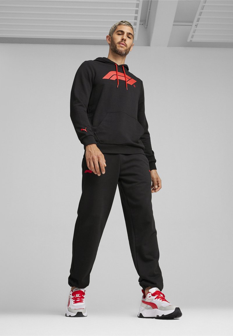 Sort hoodie med røde detaljer og logo, matchende sorte bukser og hvide sneakers med røde elementer. Materialet ser blødt og struktureret ud.