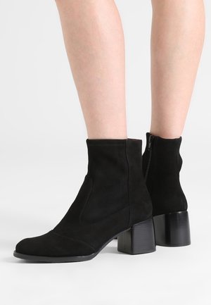 Jambes portant des bottines en suède noir avec talons blocs et fermetures éclair latérales sur fond blanc.