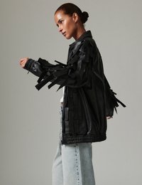 Μαύρο δερμάτινο μπουφάν με διακοσμητικά κρόσσια στα μανίκια, ελαστική μέση, μπροστινό κλείσιμο με φερμουάρ, φορεμένο πάνω από λευκή μπλούζα και ανοιχτόχρωμο τζιν.