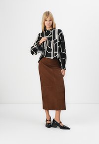 Donna bionda che indossa una blusa a strisce geometriche bianco e nero, una gonna midi marrone, tacchi neri e porta una piccola borsa nera.