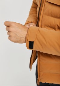 Brązowa pikowana kurtka o gładkiej fakturze. Posiada zapięcie na rzep przy mankietach i zamek błyskawiczny z przodu. Kołnierz bez zapięcia, o smukłym kroju.