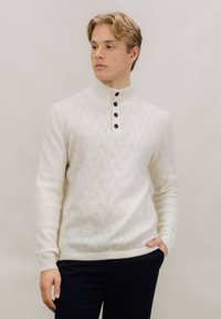 Kremfarget strikkegenser med høy krage, med et teksturert sikksakkmønster og tre mørke knapper ved halsen.