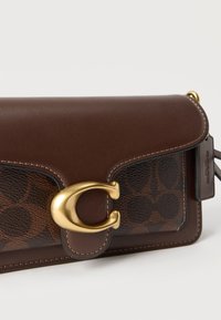 Καφέ δερμάτινη και επενδυμένη τσάντα χειρός με χρυσαφί κούμπωμα σε σχήμα "C" και ραφές στο ίδιο χρώμα, σε λευκό φόντο.