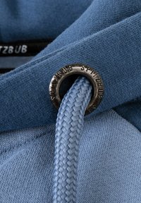 Tessuto blu con una superficie liscia, dotato di un anello metallico e di un cordoncino intrecciato di colore azzurro. Esposizione del logo su una banda elastica.