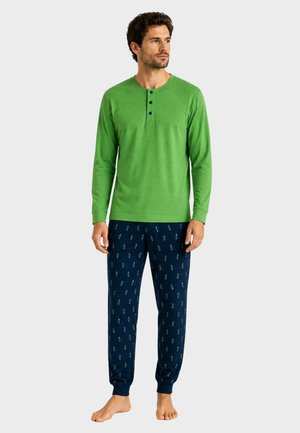 Uomo che indossa una maglietta henley verde a maniche lunghe e pantaloni da pigiama blu navy con piccoli motivi, in piedi a piedi nudi su uno sfondo uniforme.