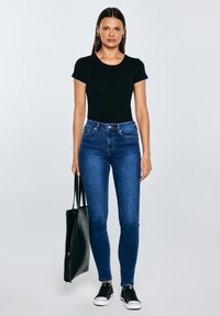 Vrouw staand, gekleed in een zwart topje met korte mouwen, donkerblauwe strakke jeans, zwarte sneakers, en met een grote zwarte shopper in haar linkerhand.