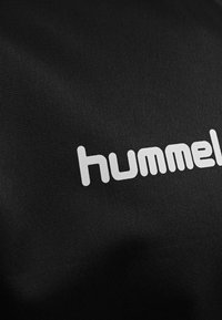 Schwarzer Stoff mit einer strukturierten Oberfläche, der ein auffälliges weißes "hummel"-Logo zeigt, das ein sauberes, modernes typografisches Design präsentiert.
