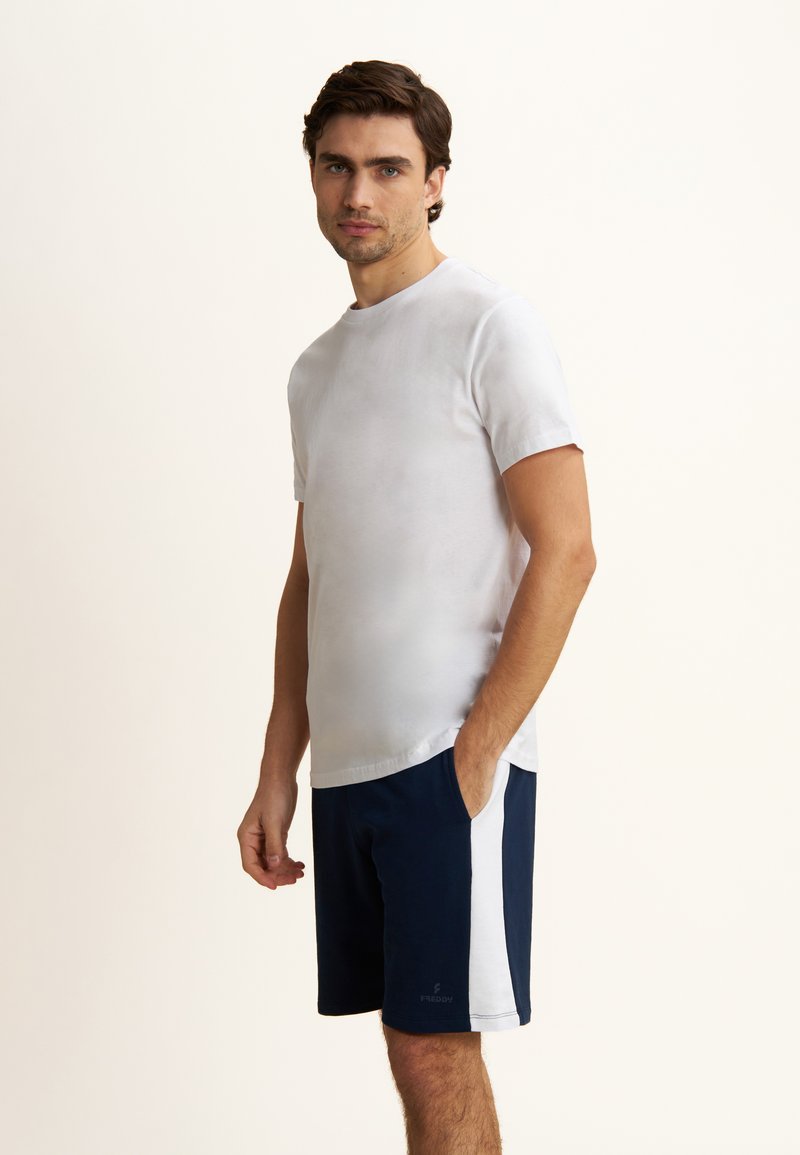 T-shirt bianco a maniche corte abbinato a pantaloni corti blu navy con strisce laterali bianche. Il tessuto appare morbido e liscio, con una vestibilità comoda.