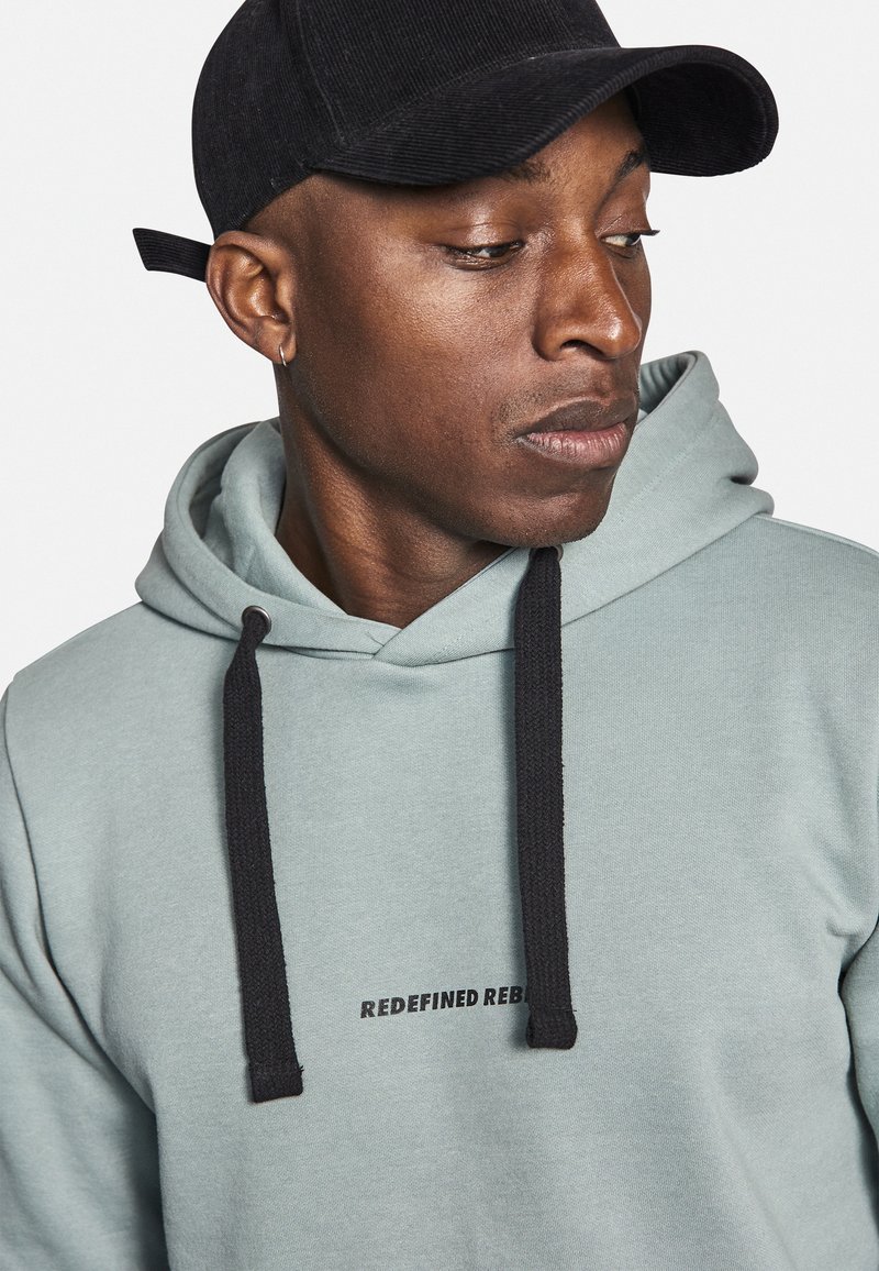 Helle grüne Kapuzenjacke mit schwarzen Kordeln, die den Text "REDEFINED REBEL" auf der Brust trägt; kombiniert mit einer schwarzen Cordmütze.