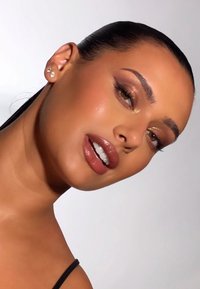 Porträt einer Person mit glatter, bronzefarbener Haut, glänzenden braunen Lippen, dezentem Augen-Make-up und Ohrsteckern, aufgenommen vor einem neutralen Hintergrund.