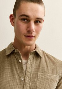 Młody mężczyzna z krótkimi włosami, ubrany w beżową koszulę z fakturą na guziki, noszącą jasną bluzkę w środku, patrzy bezpośrednio w kamerę.